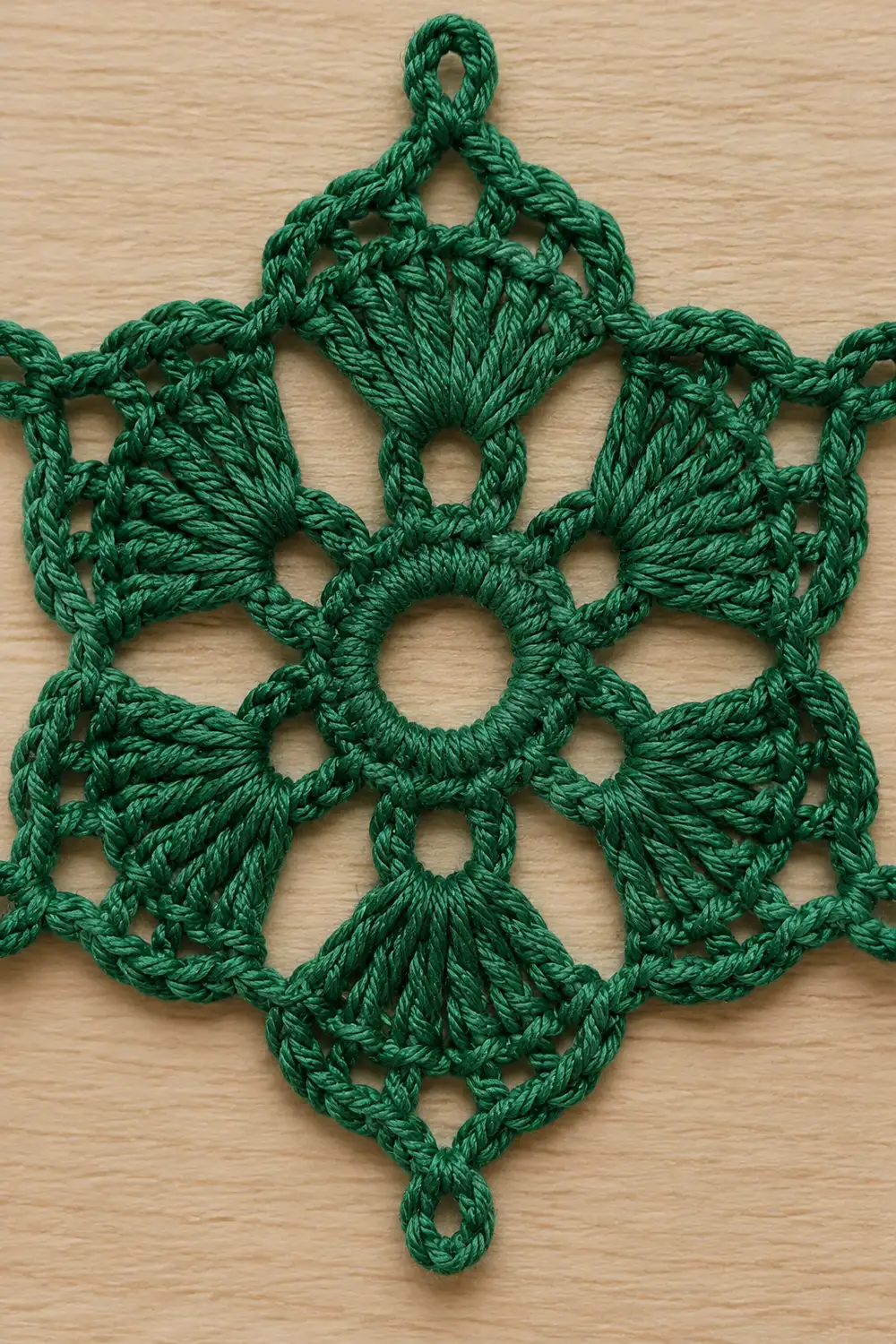 A green crocheted snowflake ornament with intricate lacework details lying flat on a light wood surface.