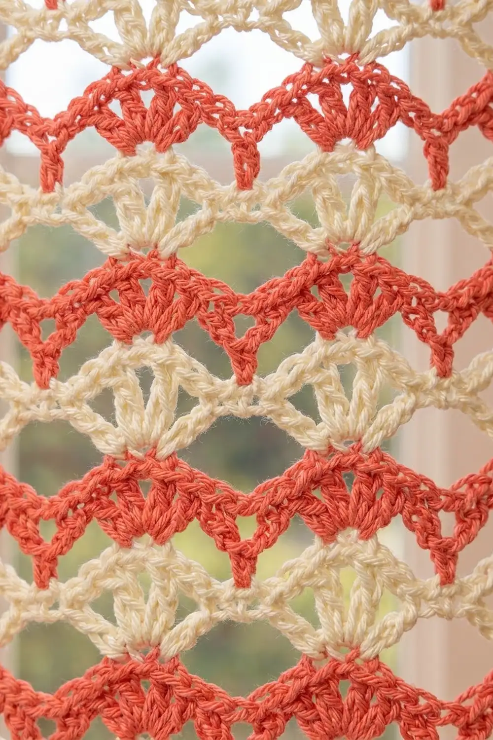 Cream and coral-red crochet lace in a repeating diamond pattern with open lacework holes against a blurred garden backdrop.