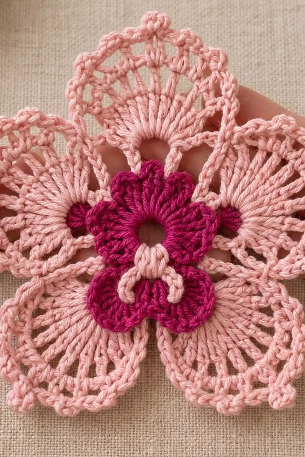 Close-up of a crocheted flower with a deep magenta center and light pink petals on cream linen fabric.