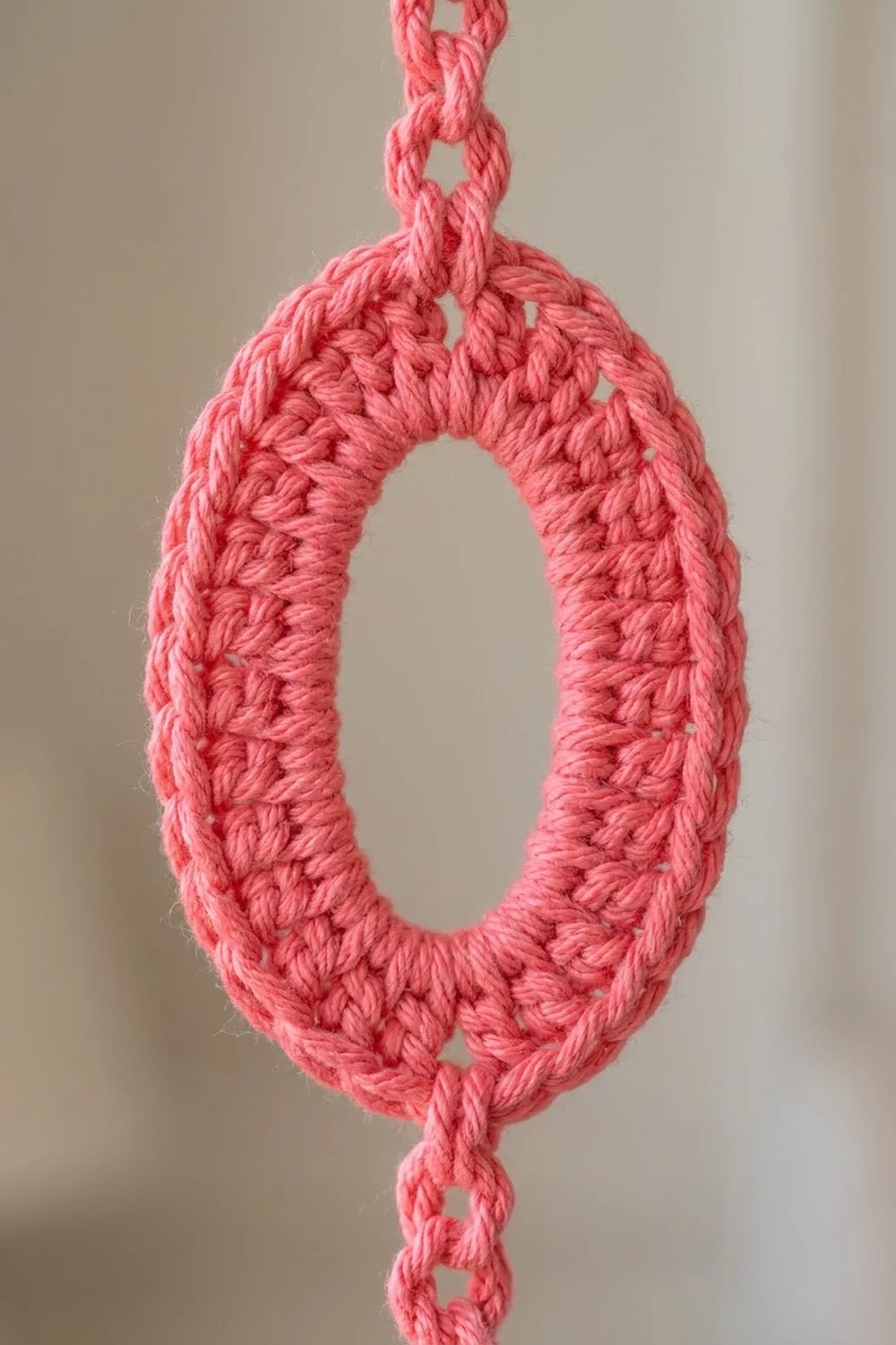 Pink crocheted loop with detailed texture stitching suspended against a neutral beige background.