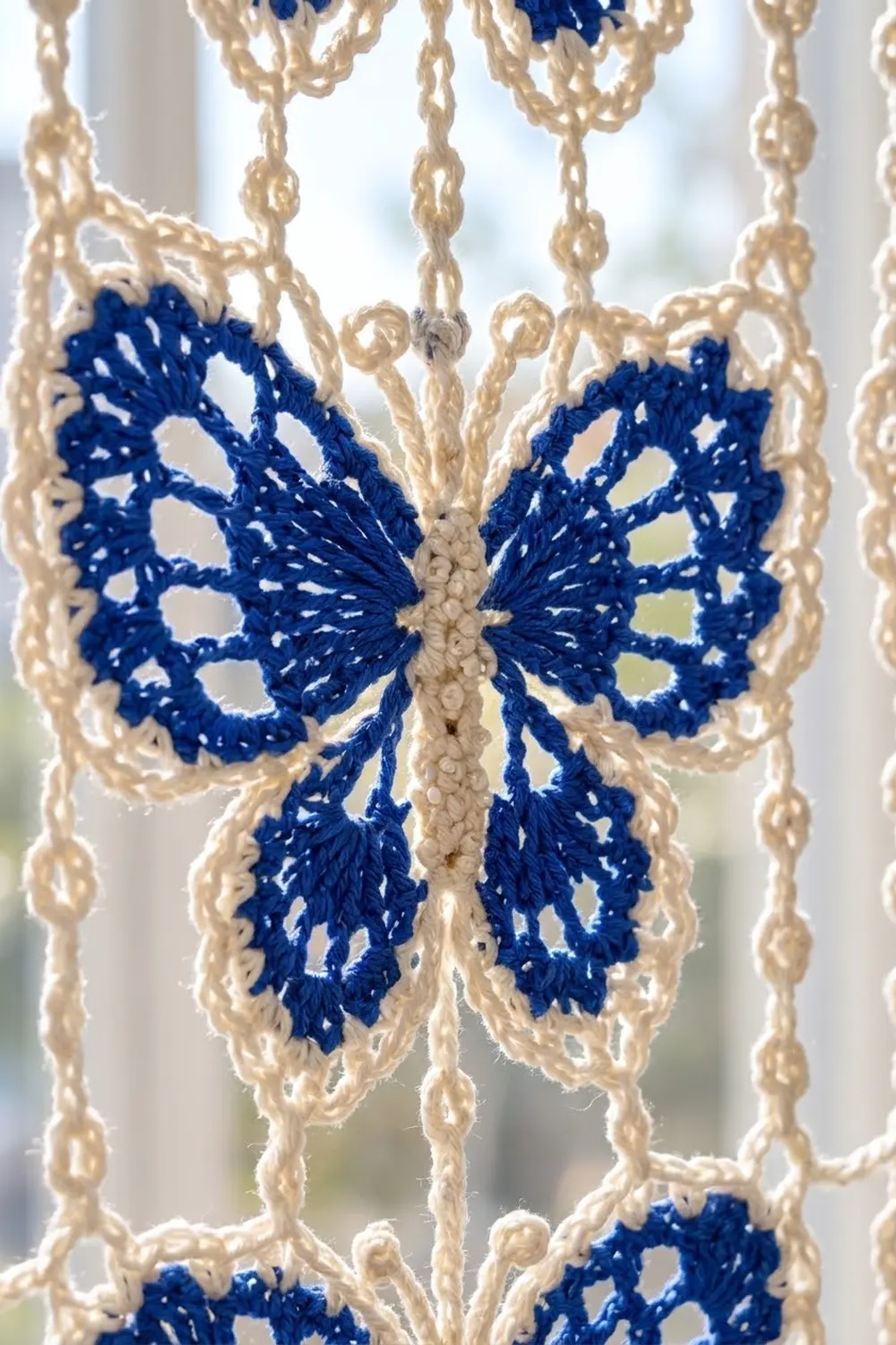 A navy and cream crocheted butterfly ornament hangs in natural light, displaying intricate lace detailing in this handmade mobile piece.