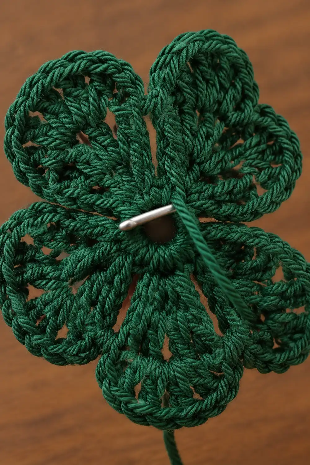 A crocheted four-leaf clover in green yarn with a silver crochet hook against a brown background.