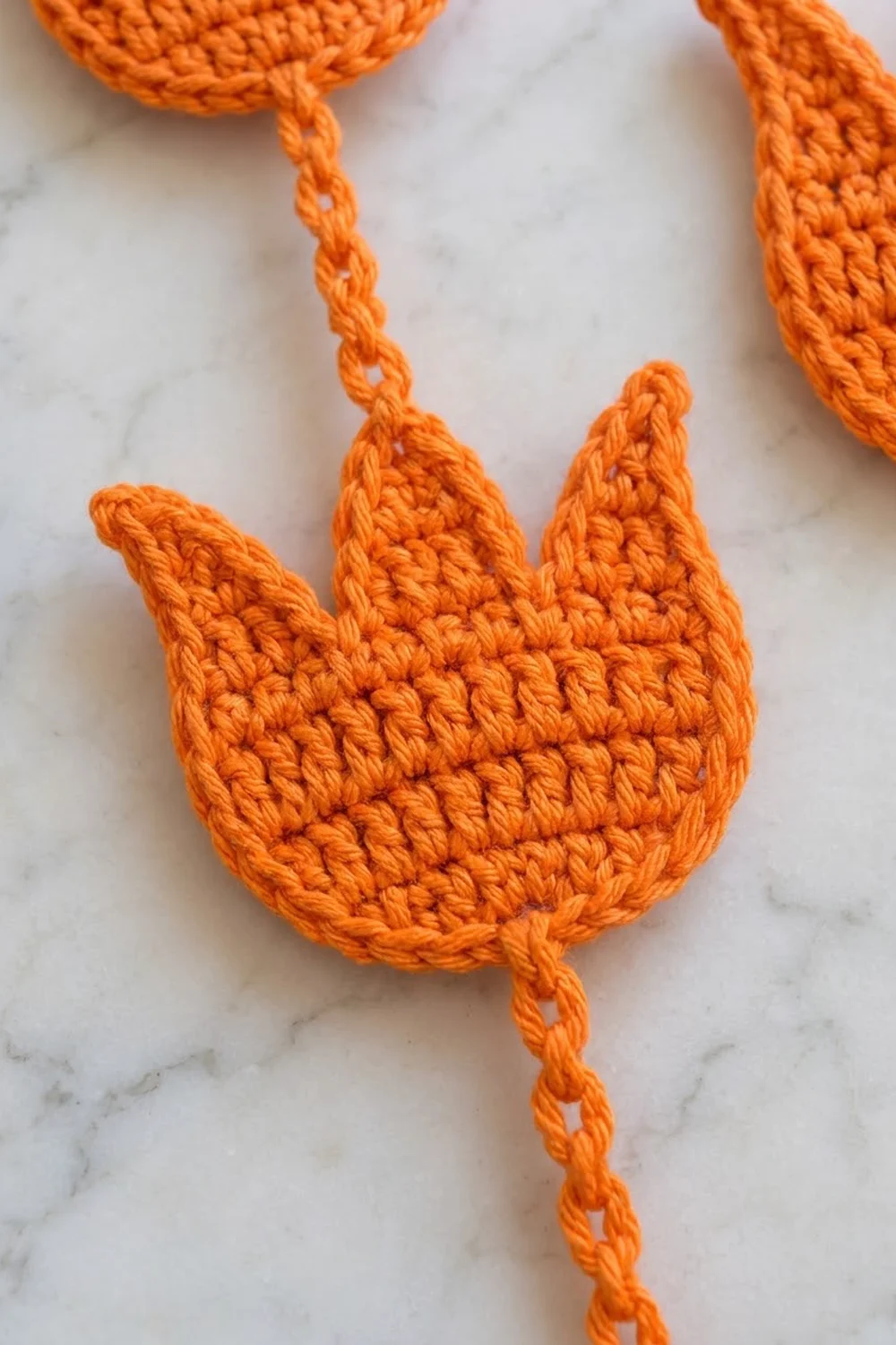 Close-up of a bright orange crocheted flower with three pointed petals on a white marble surface.