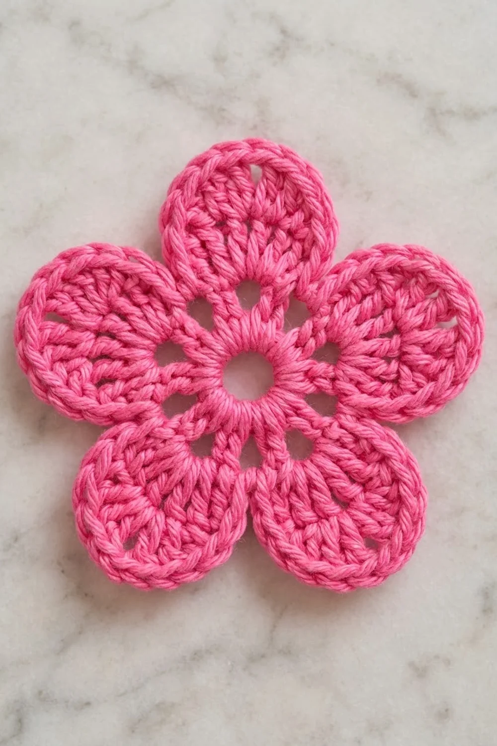 Bright pink crocheted five-petal flower with visible stitching details on a light gray marble background.