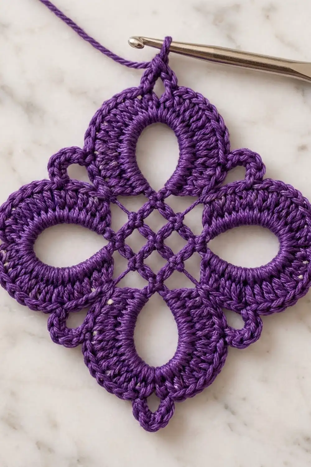 Purple crocheted ornament with delicate lace quatrefoil pattern hanging from a silver crochet hook.