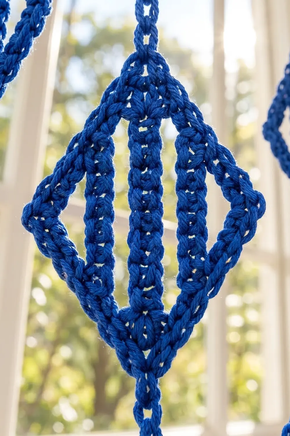 Bright blue crochet diamond with openwork pattern hanging in sunlit window overlooking garden.