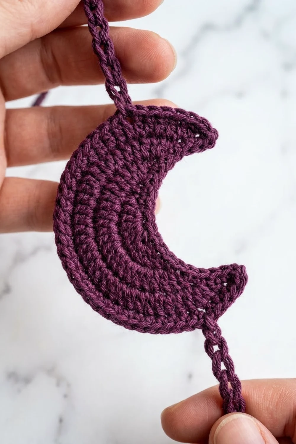 Crocheted purple curved ornament with textured stitches held between fingers against a marble-patterned background.