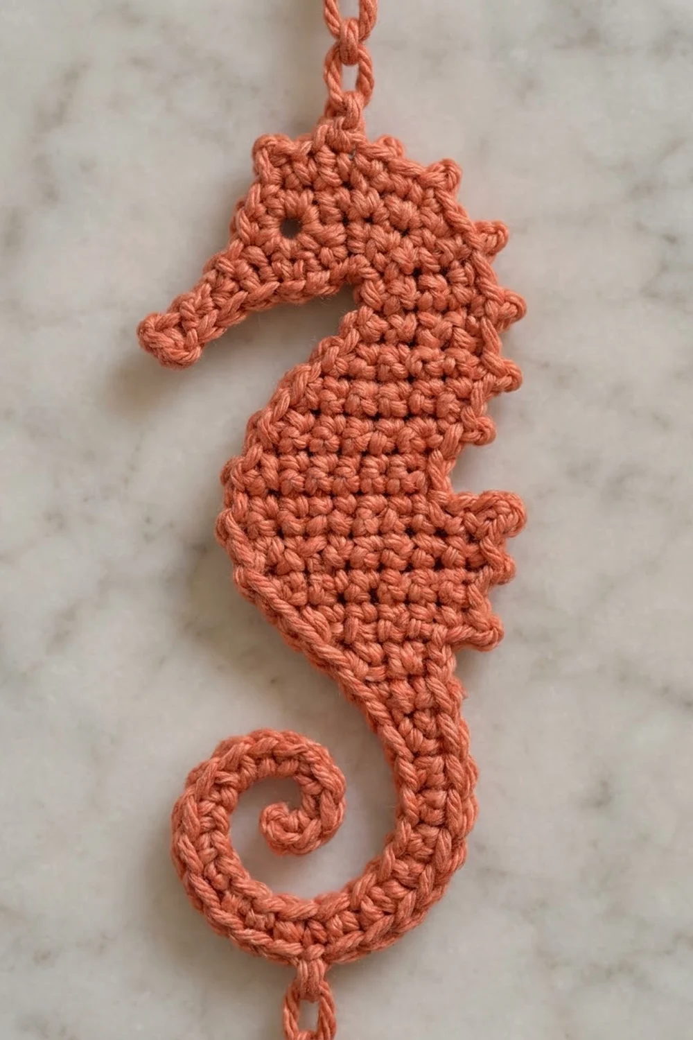 Salmon-pink crochet seahorse pendant with textured stitching hanging from a chain on marble.