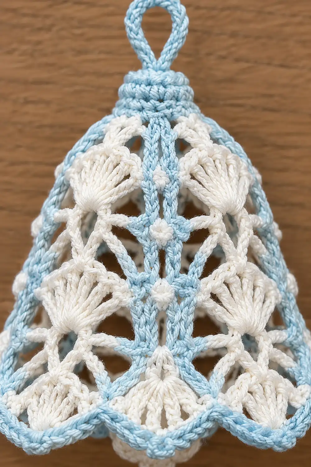 A handmade macramé bell ornament with light blue outer edges and cream-colored knotted details on a wooden surface.