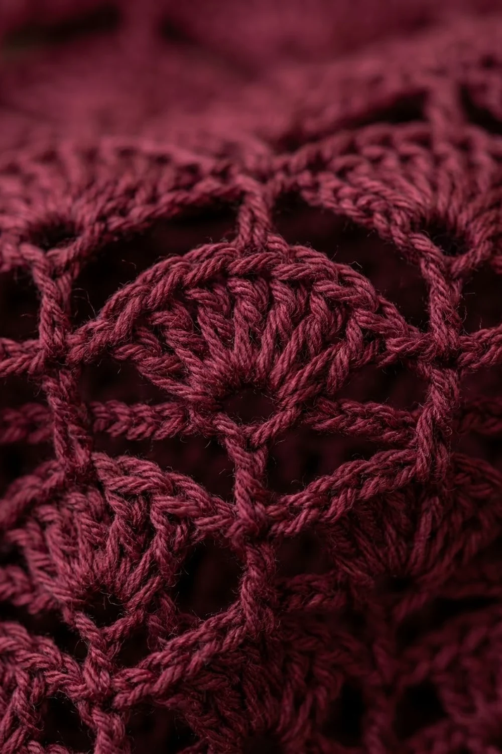 Burgundy crochet fabric displays intricate twisted stitches forming an open, lacy geometric pattern with visible yarn texture.