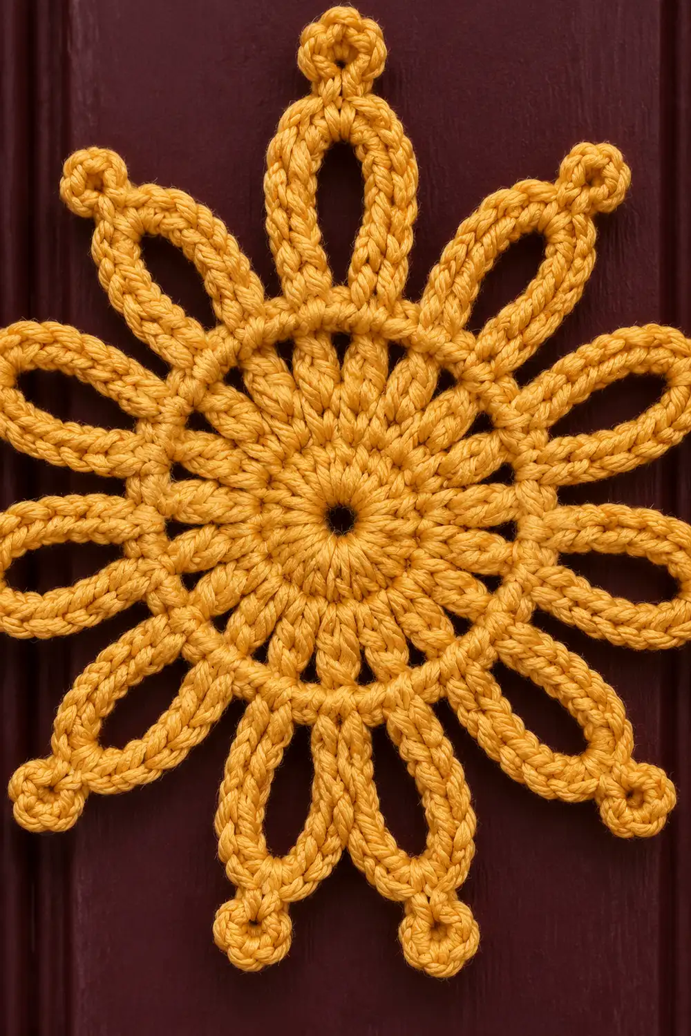 Golden crocheted snowflake ornament with twelve looped petals displayed against a deep burgundy fabric background.