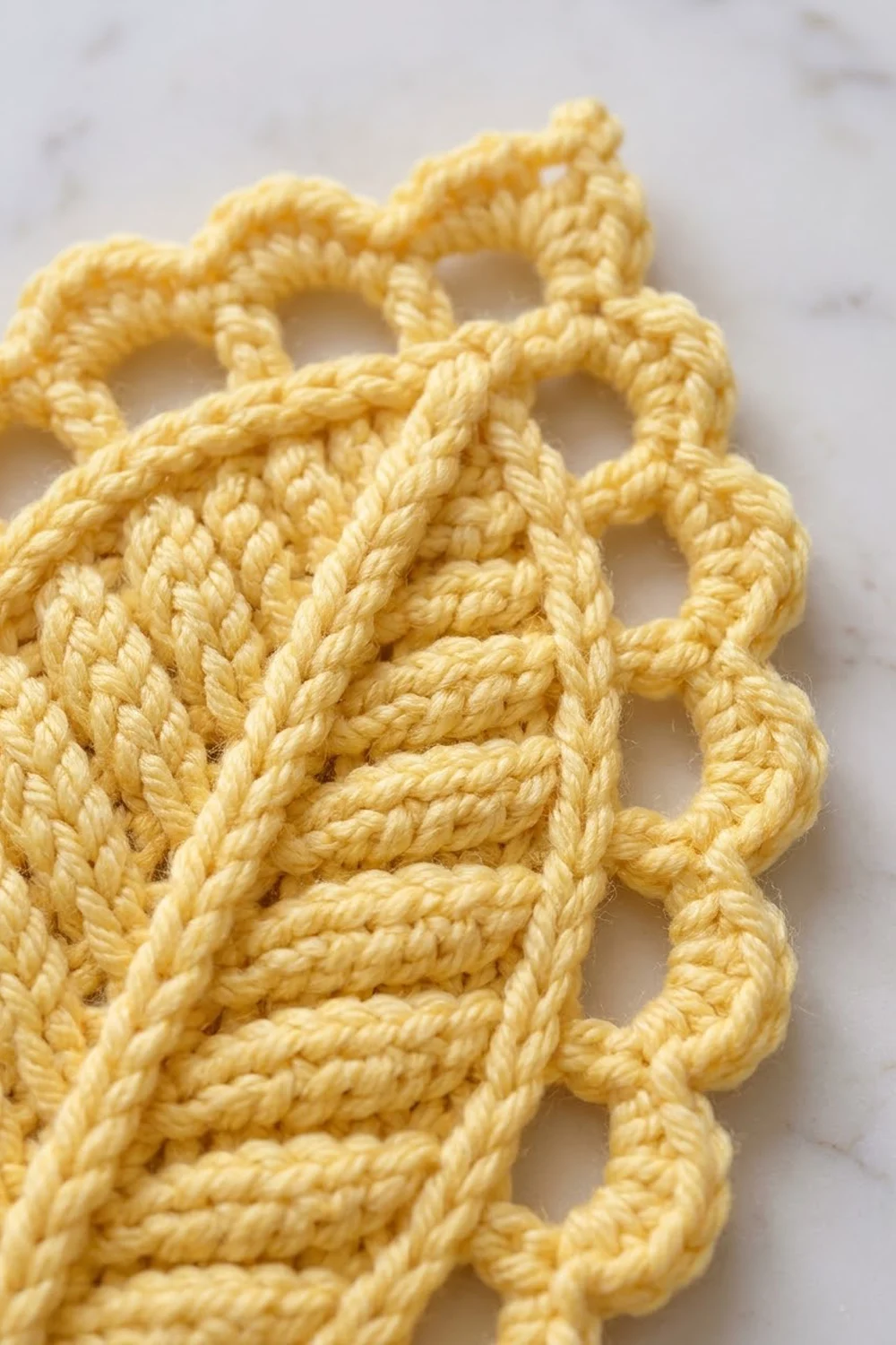 Close-up of pale golden crochet work with delicate openwork pattern and textured stitches against a light marble background.