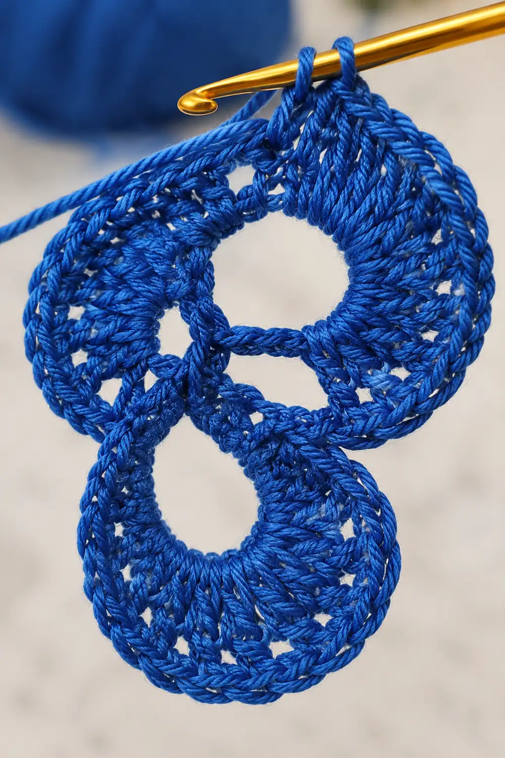 Blue crocheted lace circles hang from a golden crochet hook against a blurred background.
