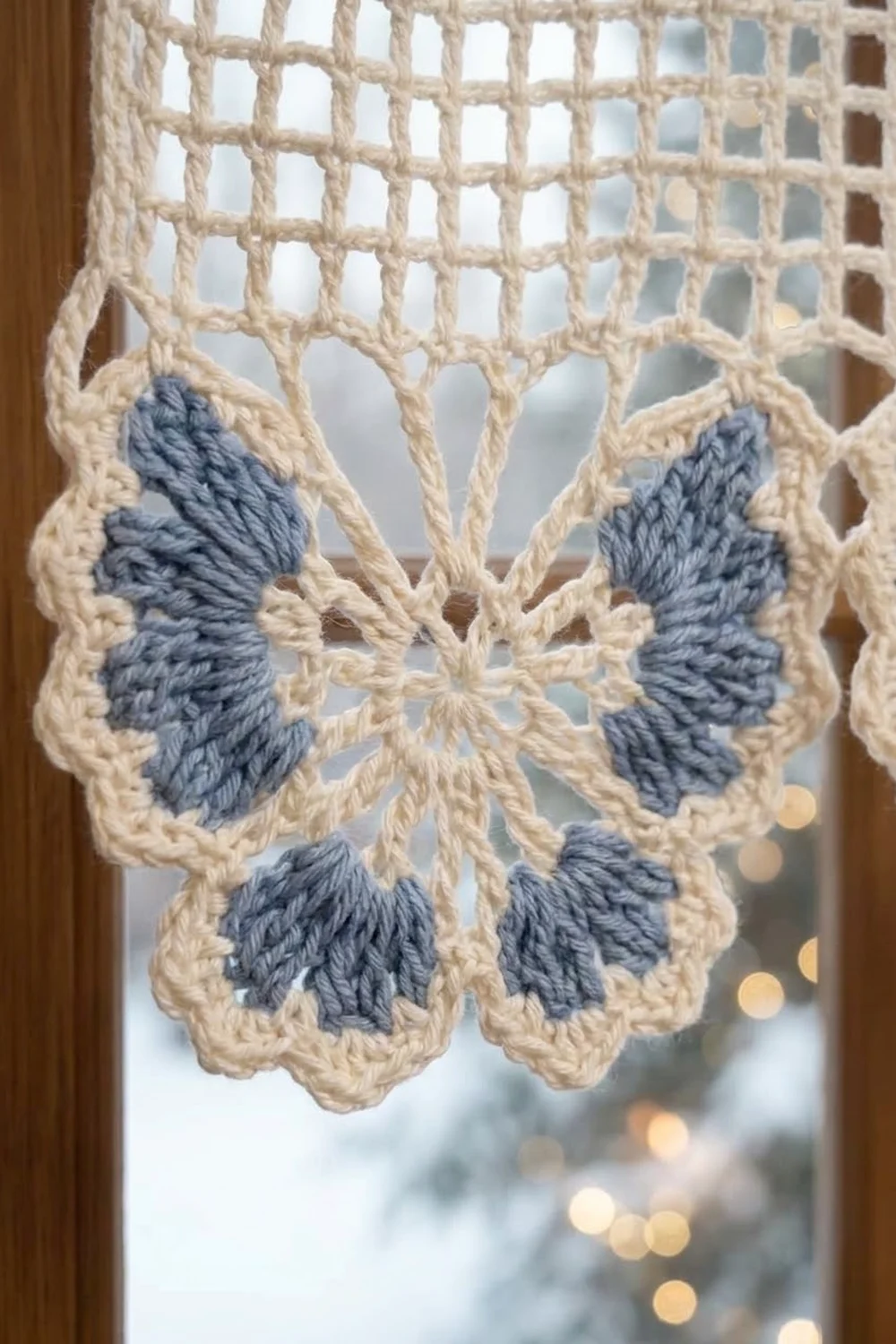 Crocheted butterfly ornament with cream lattice wings and blue-gray details, hanging in a window against blurred bokeh lights.