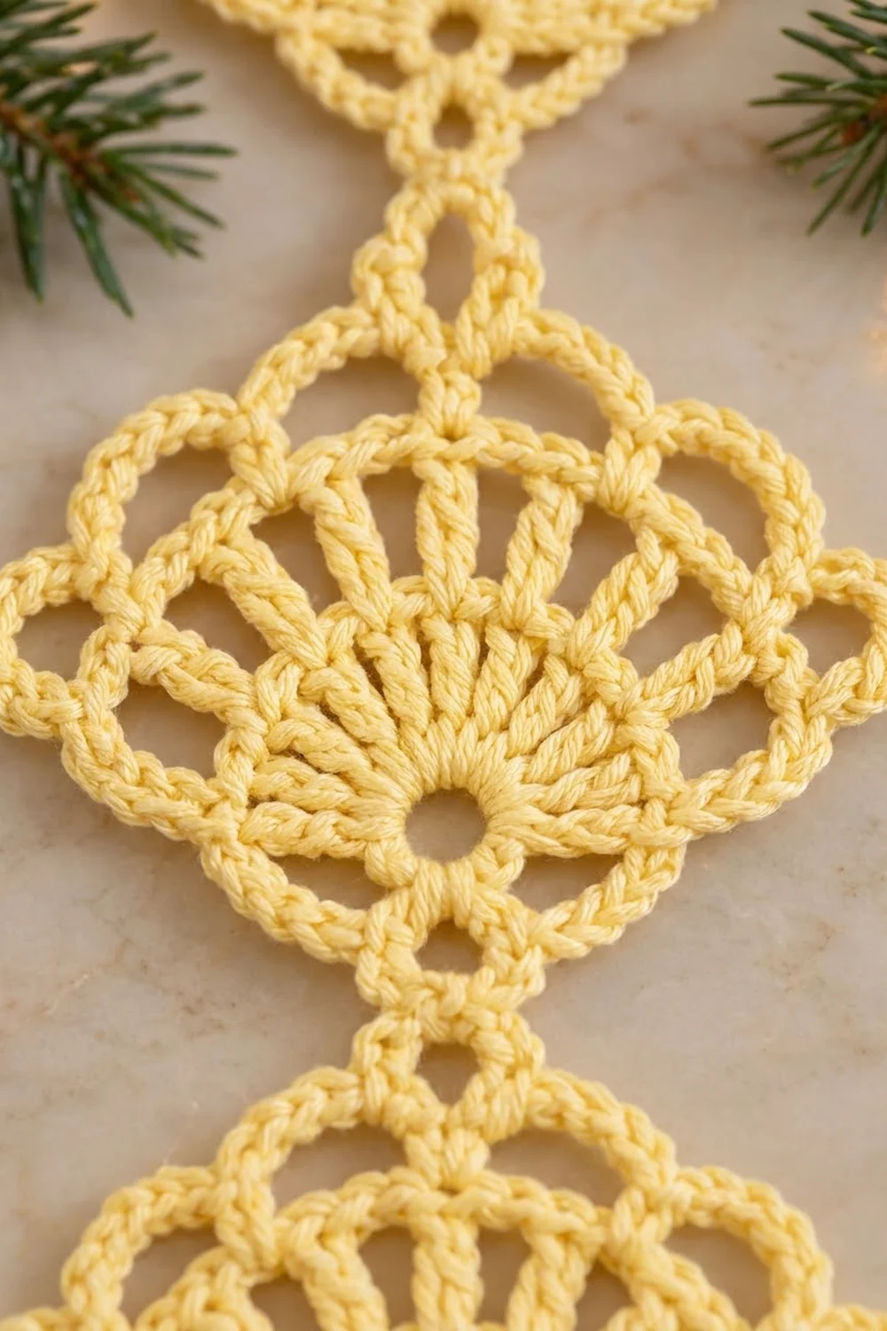 Intricate pale yellow crocheted snowflake ornament with openwork lace design displayed against neutral beige with fresh evergreen branches.