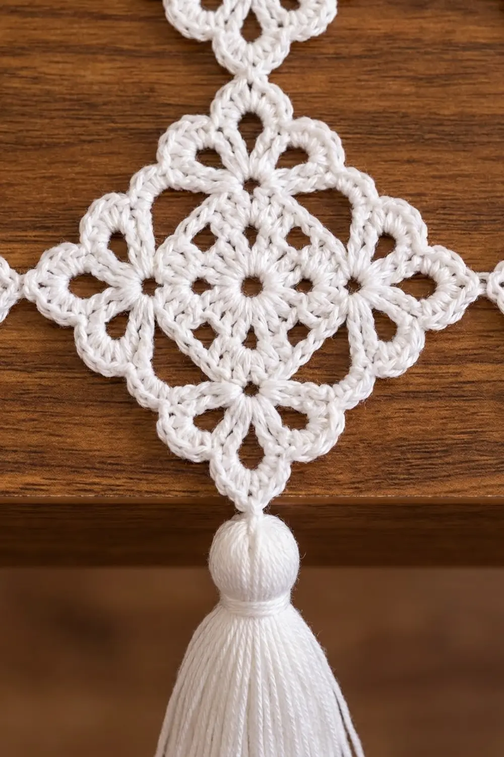Delicate white crochet ornament with intricate lace pattern and dangling tassel against warm wooden background.