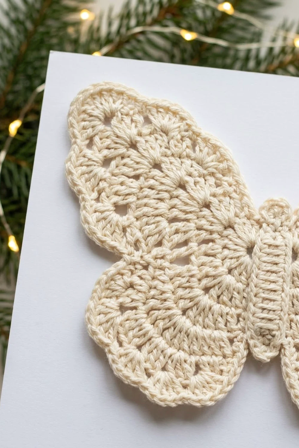 Cream crochet mitten with open stitch pattern on white card against evergreen branches and warm golden lights.