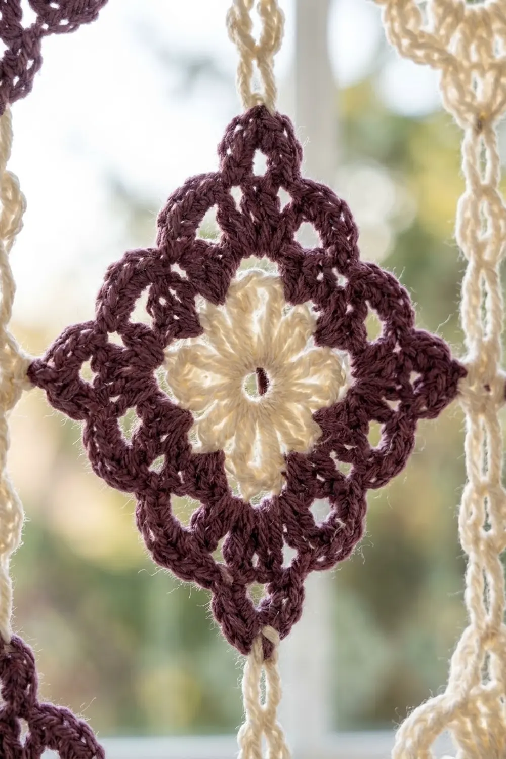 Crocheted diamond ornament in deep purple with cream floral center, hung from beige cords against a blurred garden backdrop.