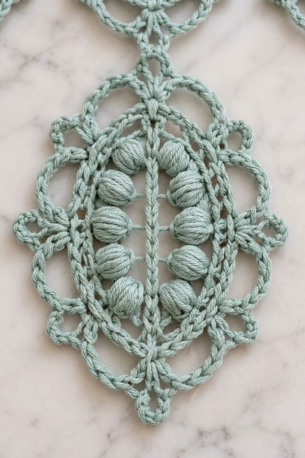 Pale sage green crocheted ornament with wrapped knots arranged symmetrically on a light marble background.