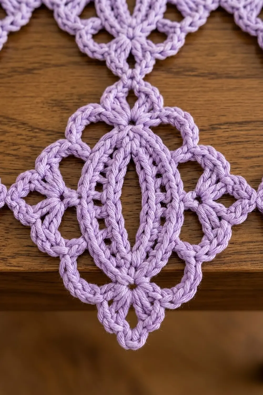 Delicate lavender crochet lace with geometric openwork motifs displayed on warm wooden surface.