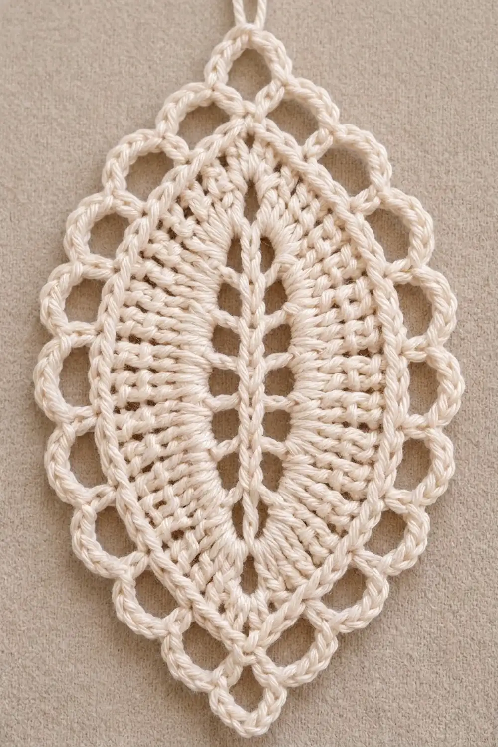 Cream-colored crocheted ornament with intricate lacy leaf pattern and twisted rope border, hanging against a beige wall.