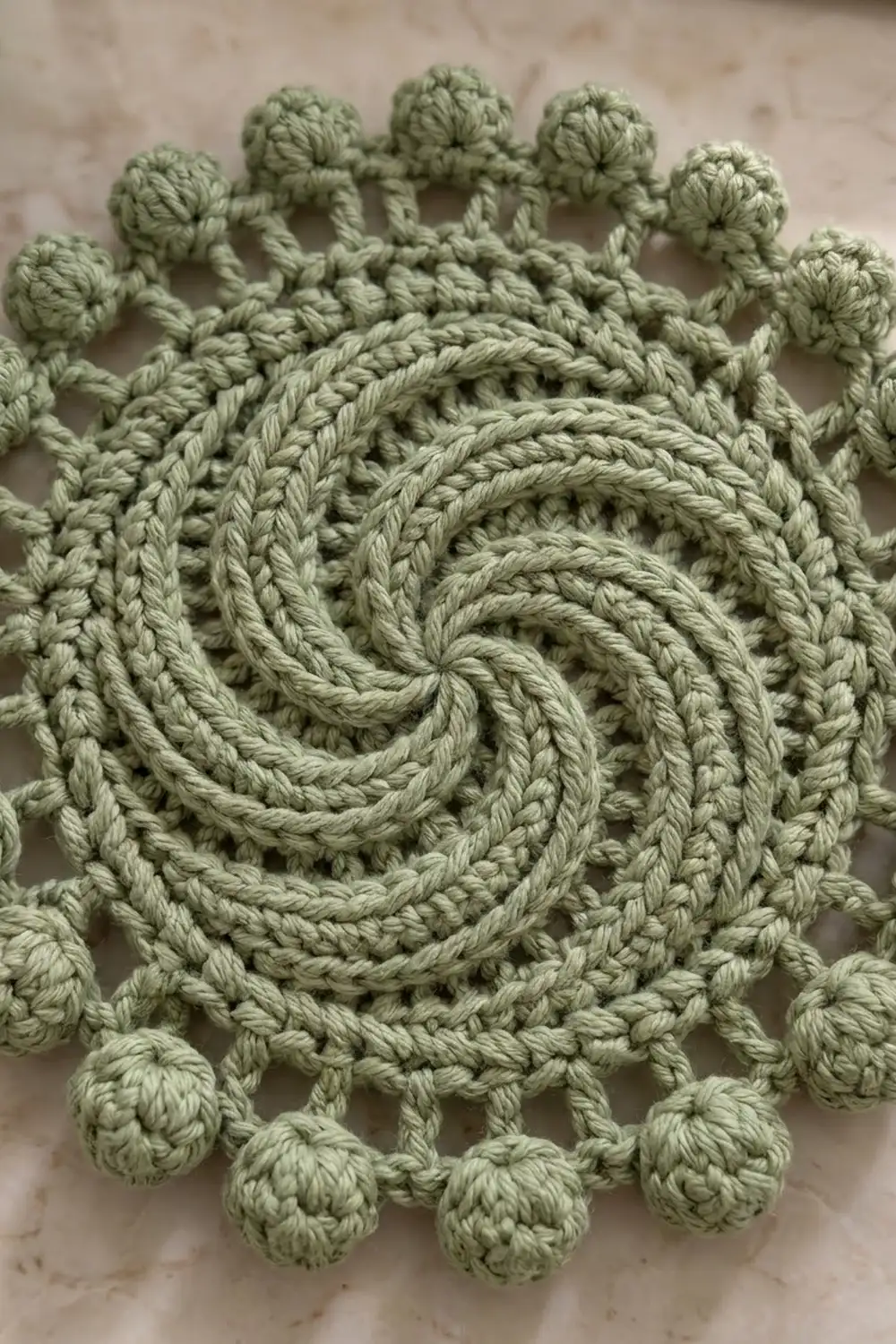 Sage green crochet motif featuring a spiral center and decorative bobble-stitch border on a neutral background.