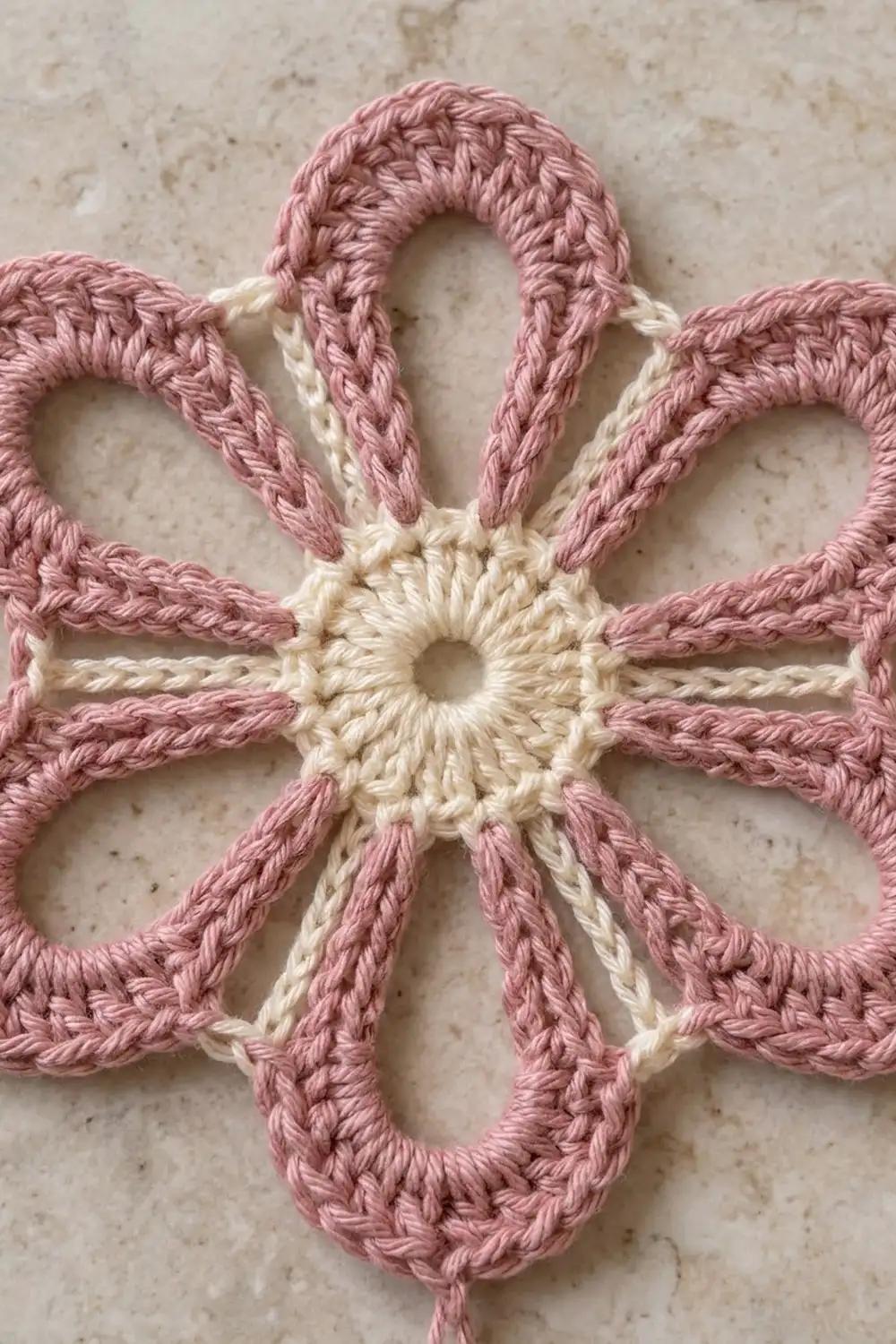 Handmade crocheted flower with mauve petals and cream center, showing intricate stitchwork on a speckled background.