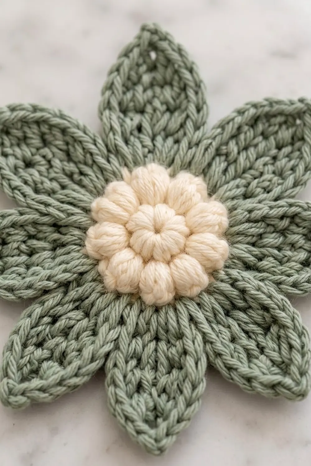 Close-up of handmade crochet flower with sage green petals, cream bobble center, and visible yarn stitch texture.