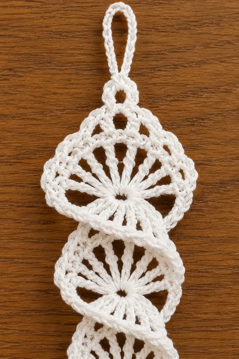 White crocheted macramé ornament with three circular medallions and loop hanging against wooden background.