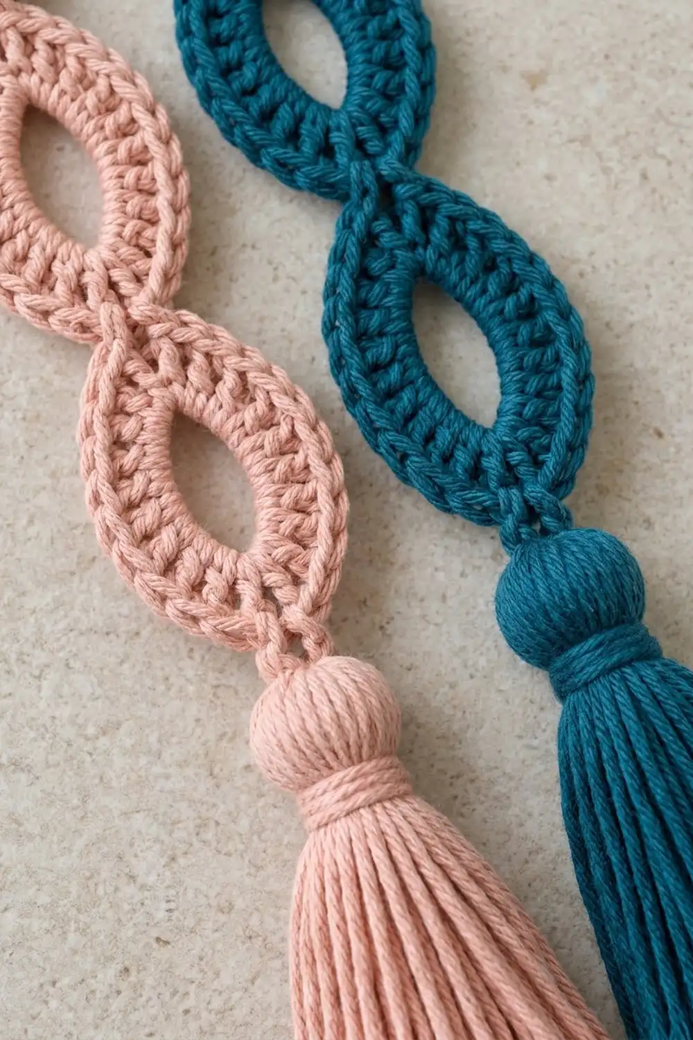 Two crocheted decorative cords in pink and teal with braided texture and bobble ends on a cream surface.
