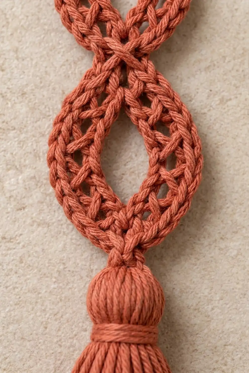 Rust-colored crocheted rope with openwork lace pattern, ending in a knotted tassel on cream fabric background.