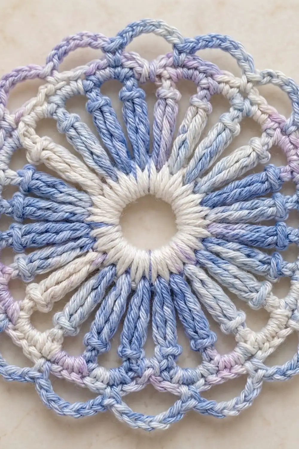 Crocheted circular doily with radiating petals in cream, blue, and lavender yarn on neutral background.