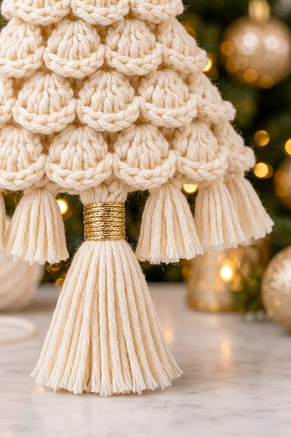 Cream crocheted Christmas tree with scalloped rows and gold metallic bow detail, holiday ornaments blurred in background.