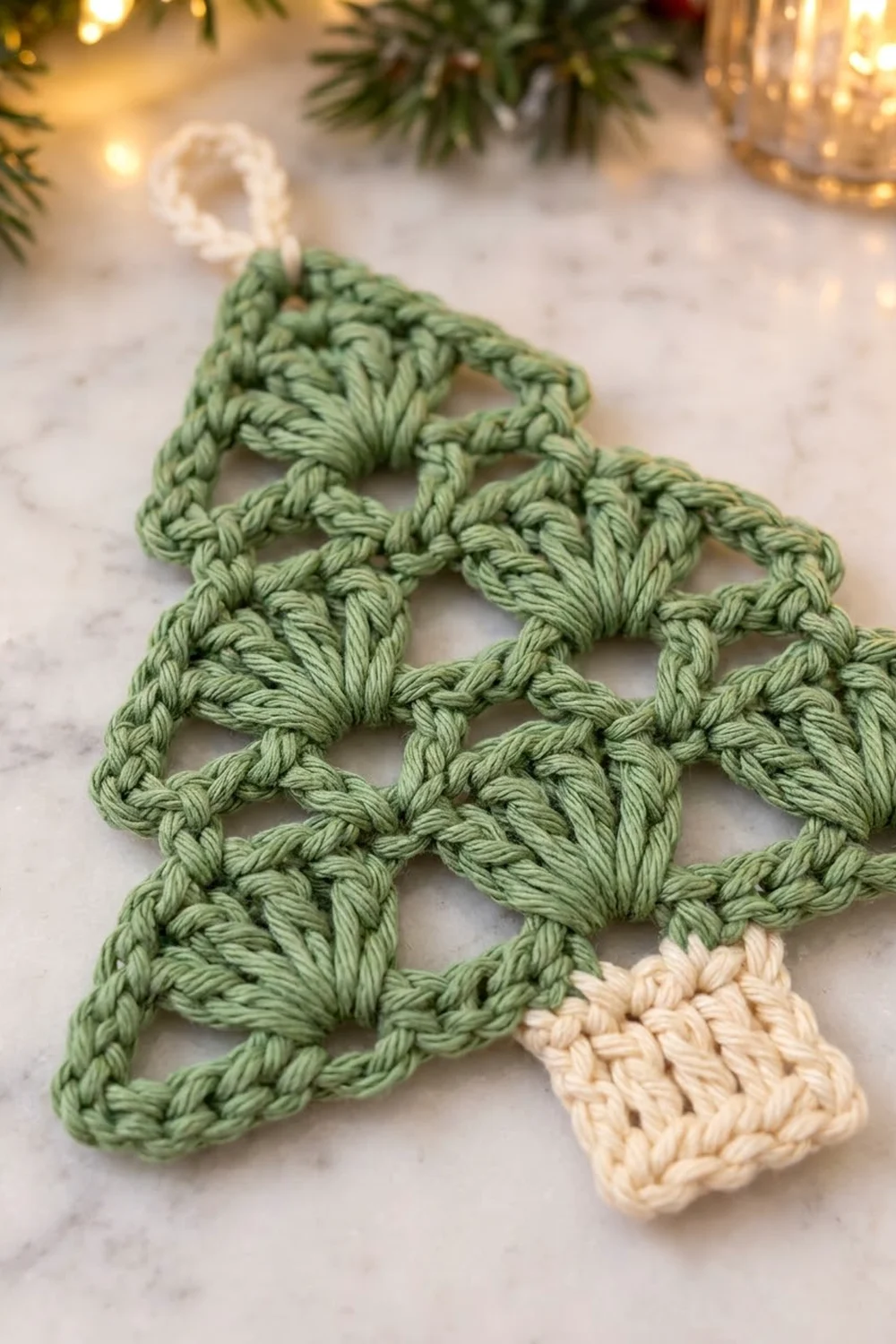 Crocheted evergreen ornament in sage green with cream accents, displayed on marble surrounded by holiday greenery and golden lights.