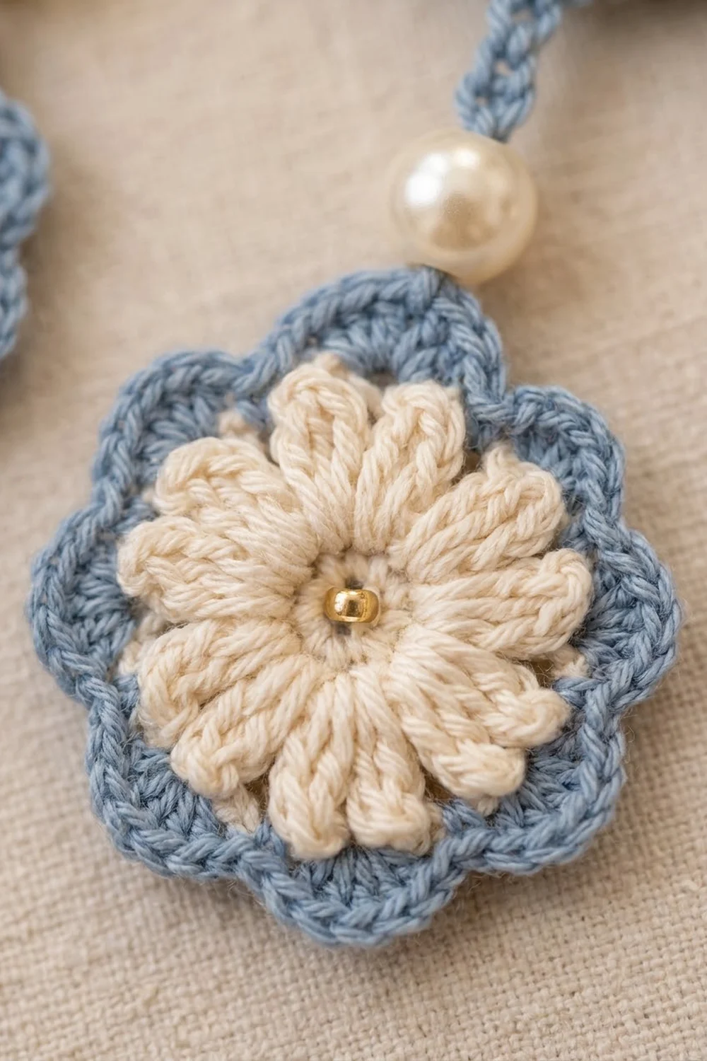 Crocheted flower with cream petals and blue-gray backing, gold bead center, attached cord, pearl bead detail on natural fabric.