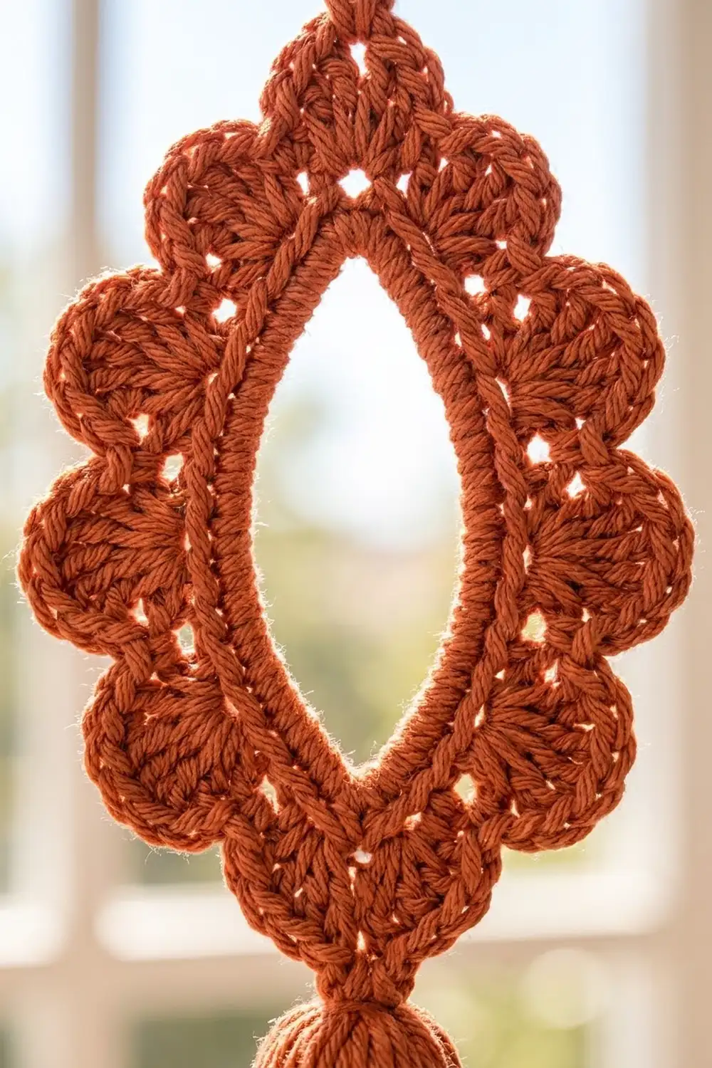 Burnt orange crocheted ornament with openwork pattern backlit against a soft, blurred window background.