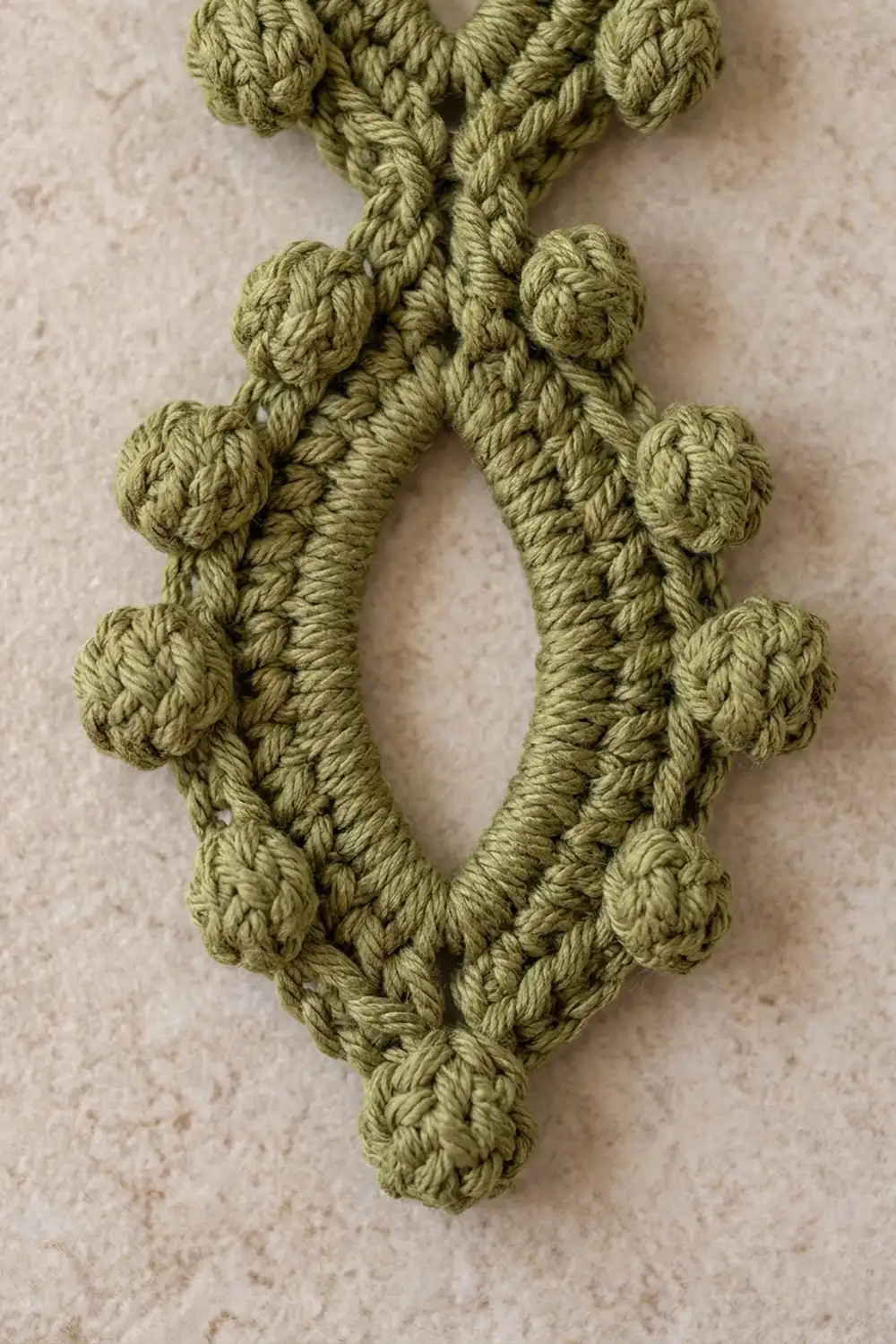 Sage green crocheted fabric with textured bobble stitches and twisted cord details on beige background.