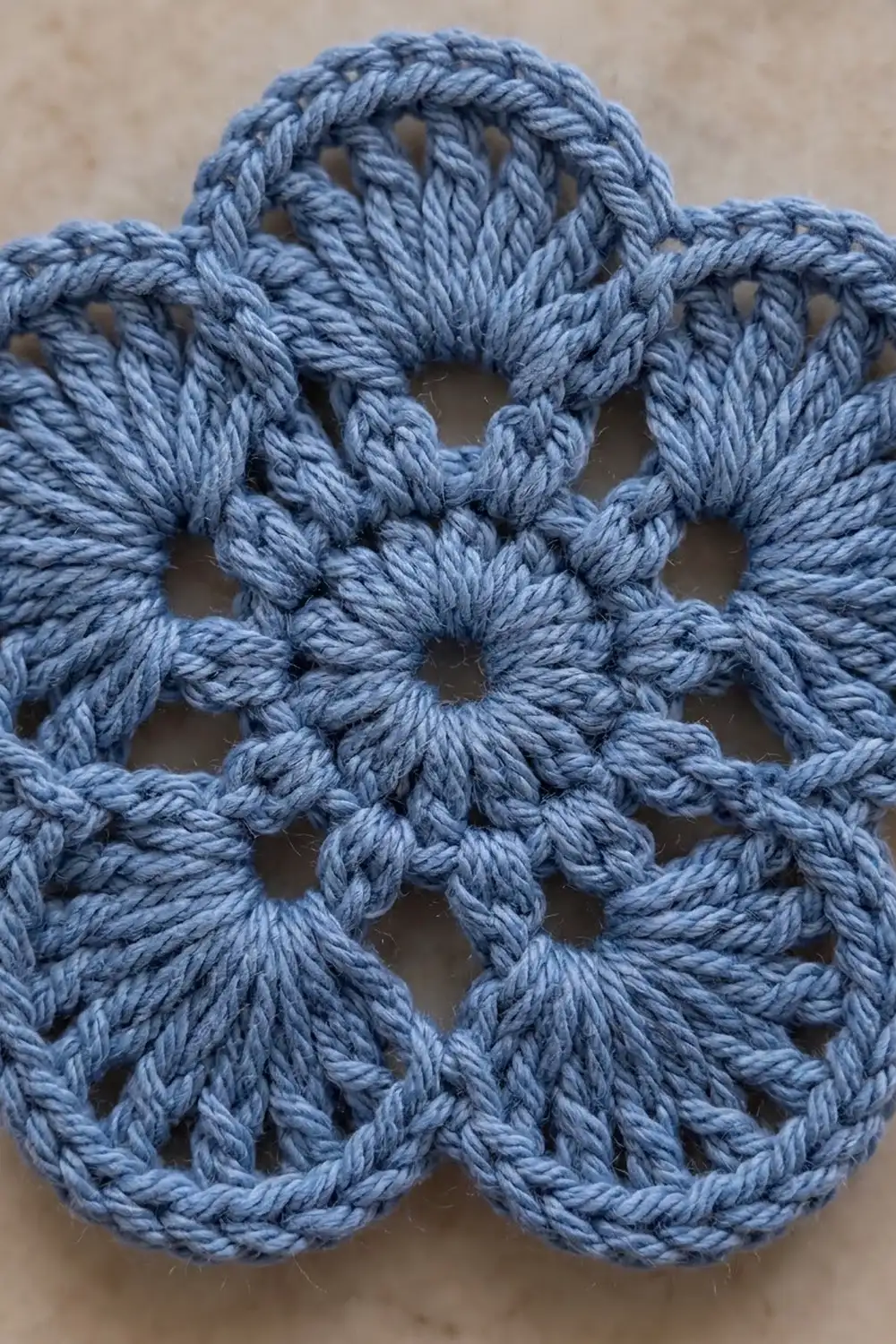 Crocheted blue-gray yarn with openwork flower motifs forming a delicate lace pattern against a soft beige background.