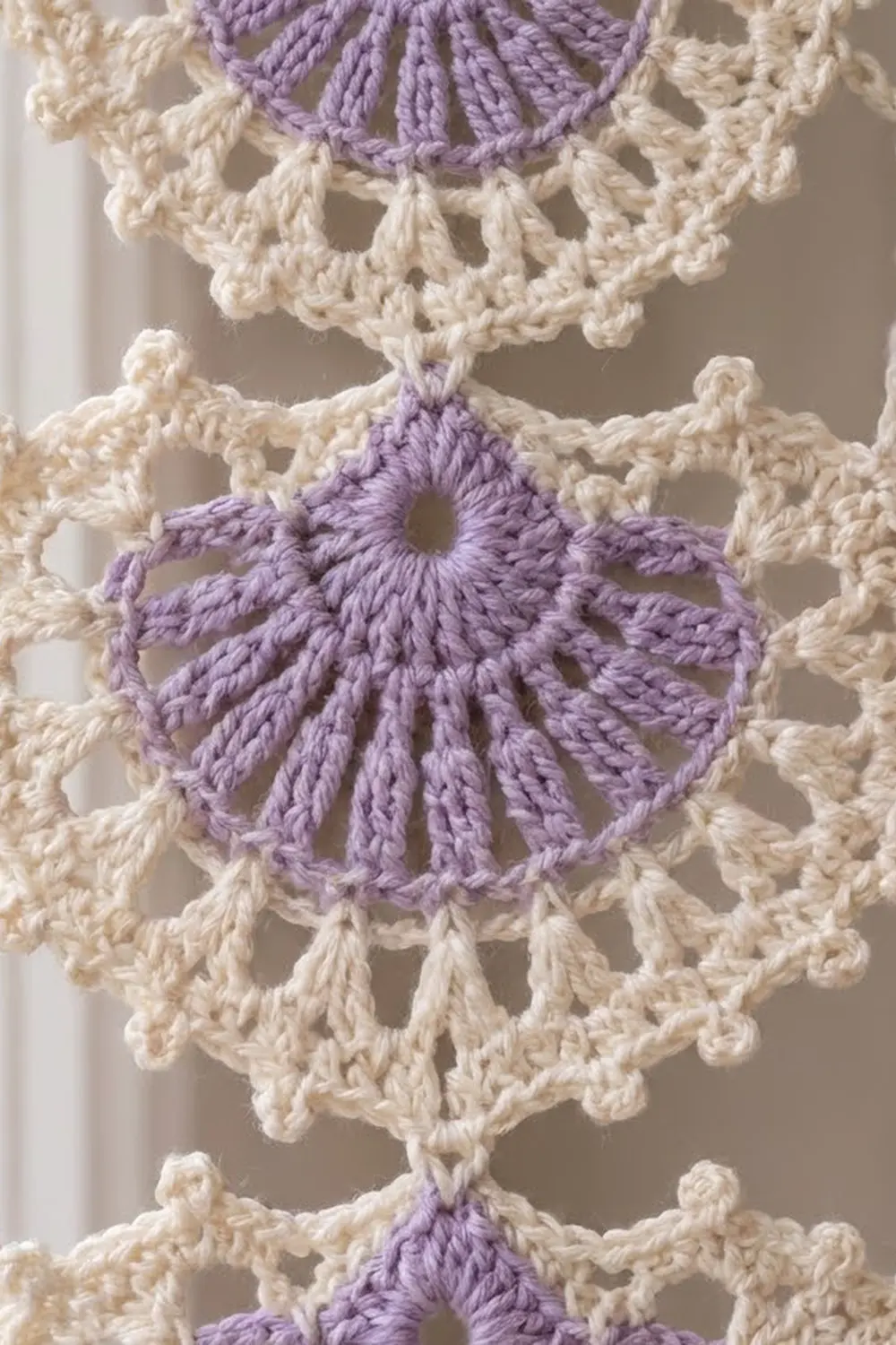 Close-up of purple and cream crocheted textile with dimensional flower medallions and delicate geometric stitch patterns.