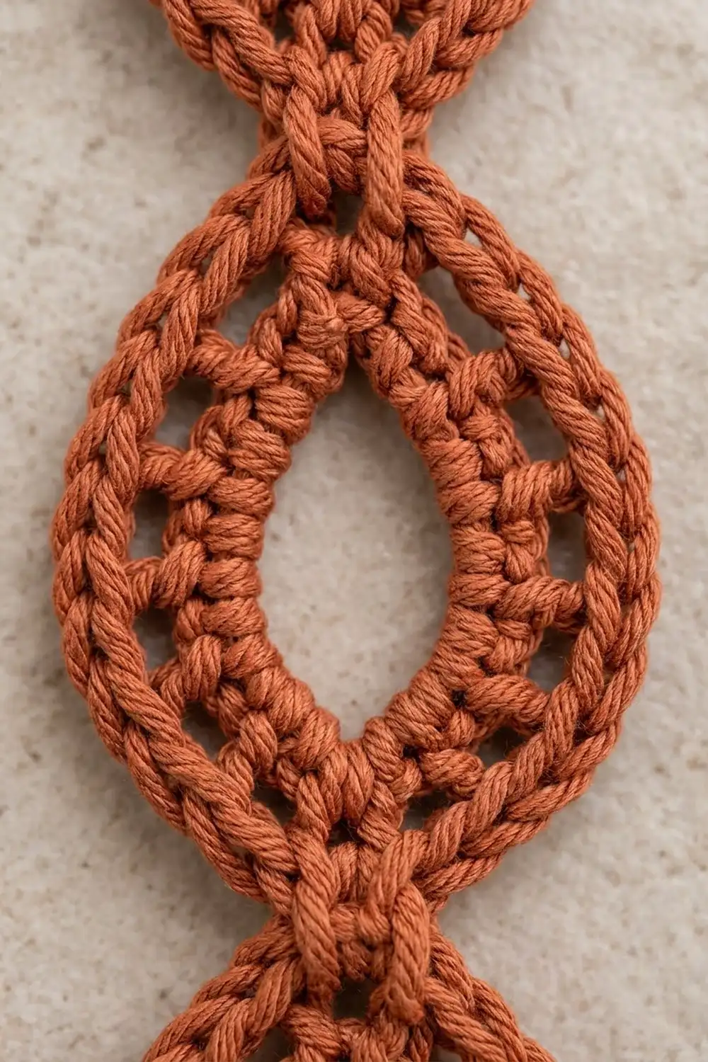 Terracotta-colored knotted macramé pendant with open teardrop design on neutral linen background.