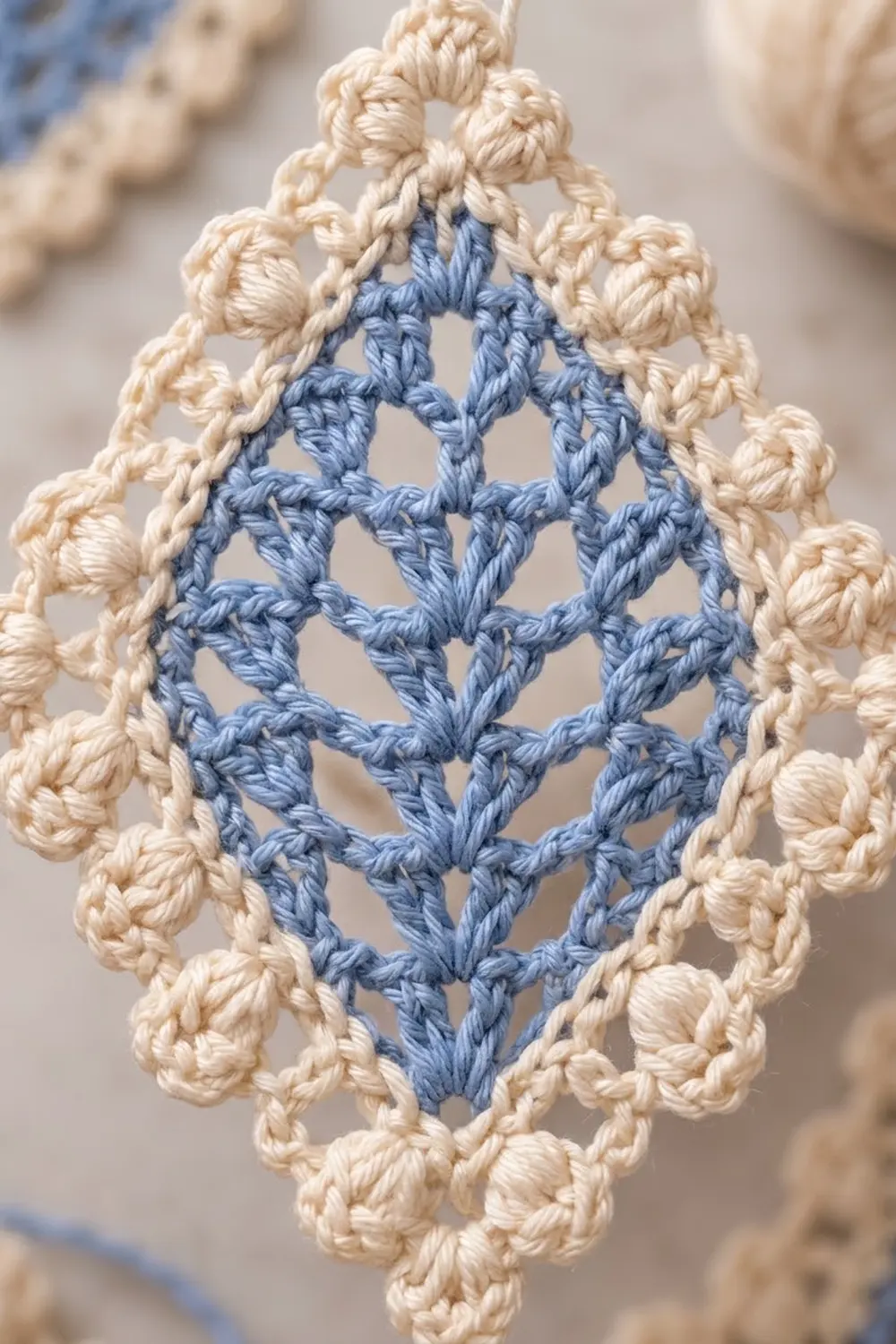 Close-up of a crocheted lace ornament with a blue diamond center and cream bobble border on a soft white background.