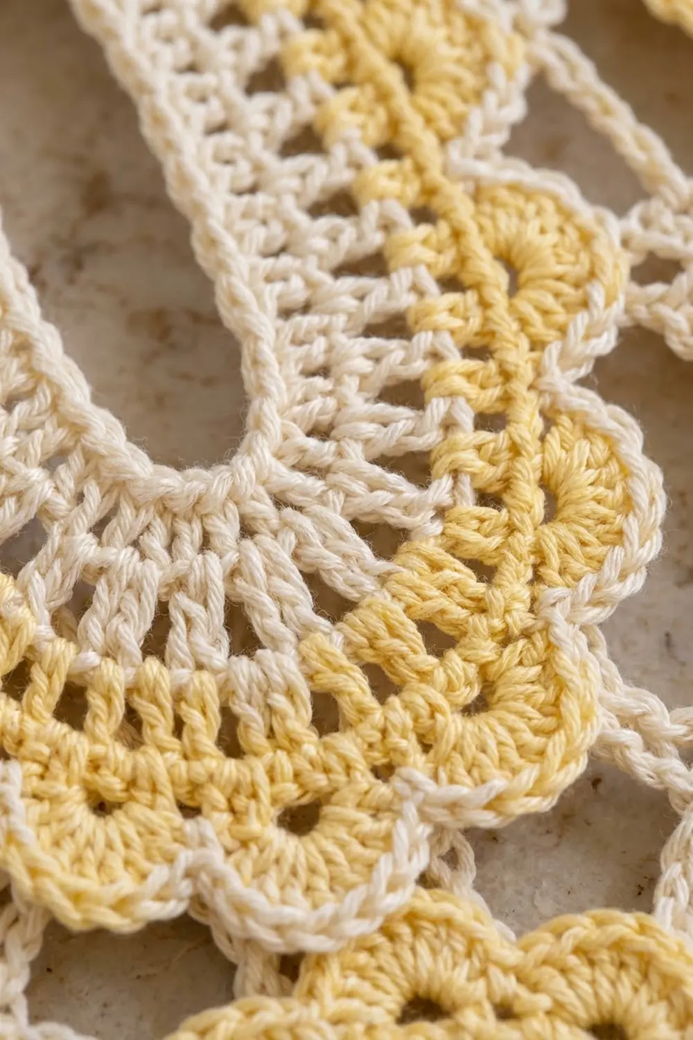 Close-up detail of cream and pale yellow crocheted lace showing intricate openwork stitching on a neutral beige background.