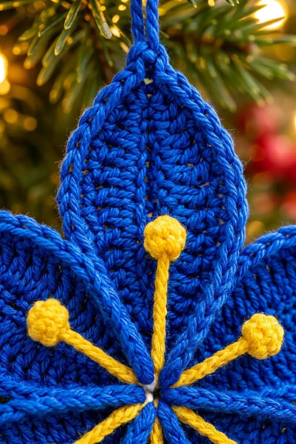 Bright blue crocheted teardrop ornament with six yellow bobbles, hanging from an evergreen branch with warm bokeh lights.