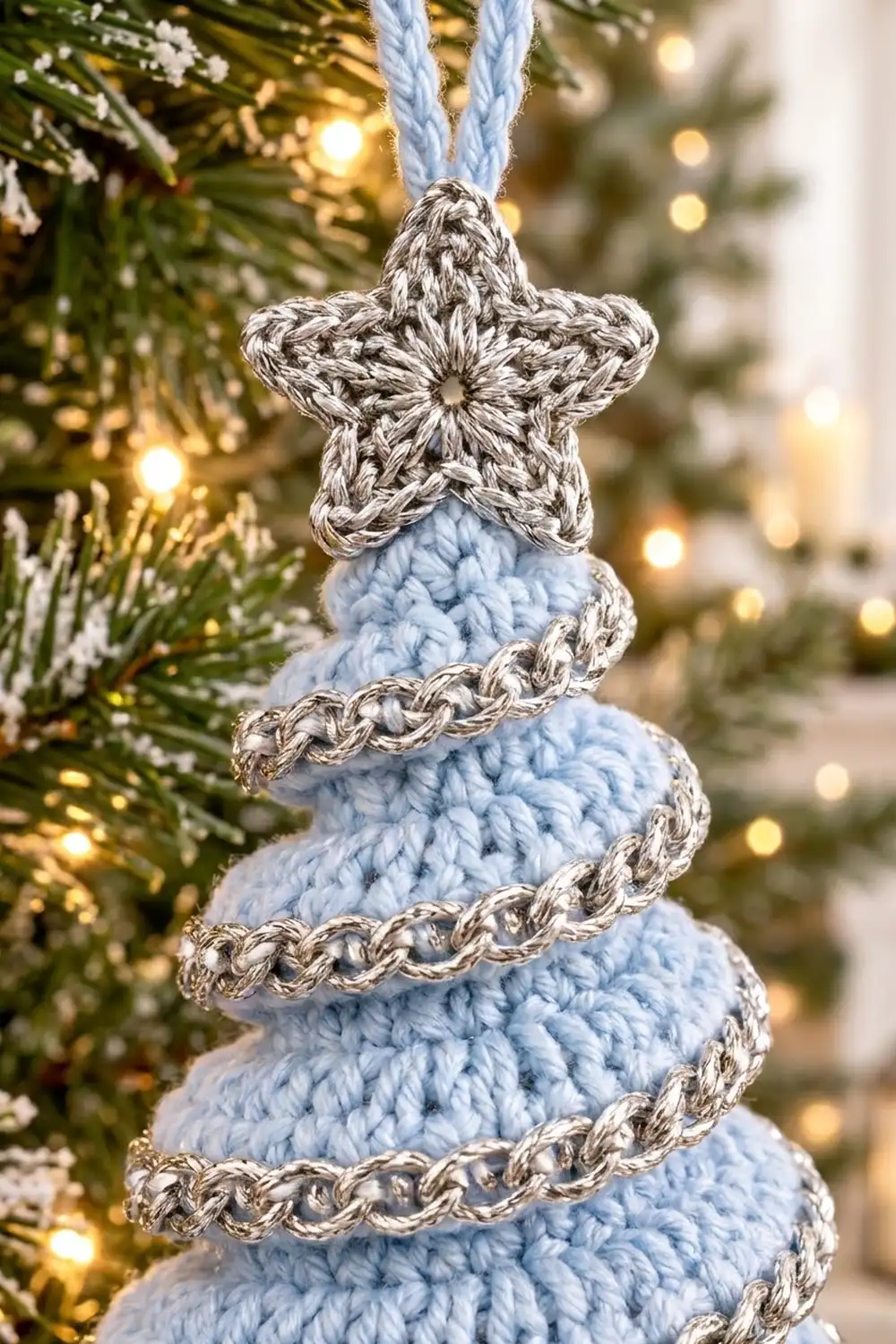 Pale blue crocheted Christmas tree ornament with silver-trimmed swirls and marled star, hanging among frosted evergreen branches with golden lights.