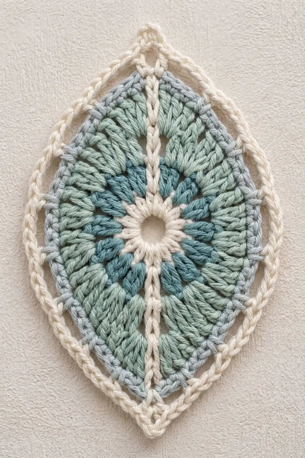 Cream-bordered crocheted oval with geometric blue-green stitched pattern and center hole mounted on white textured wall.