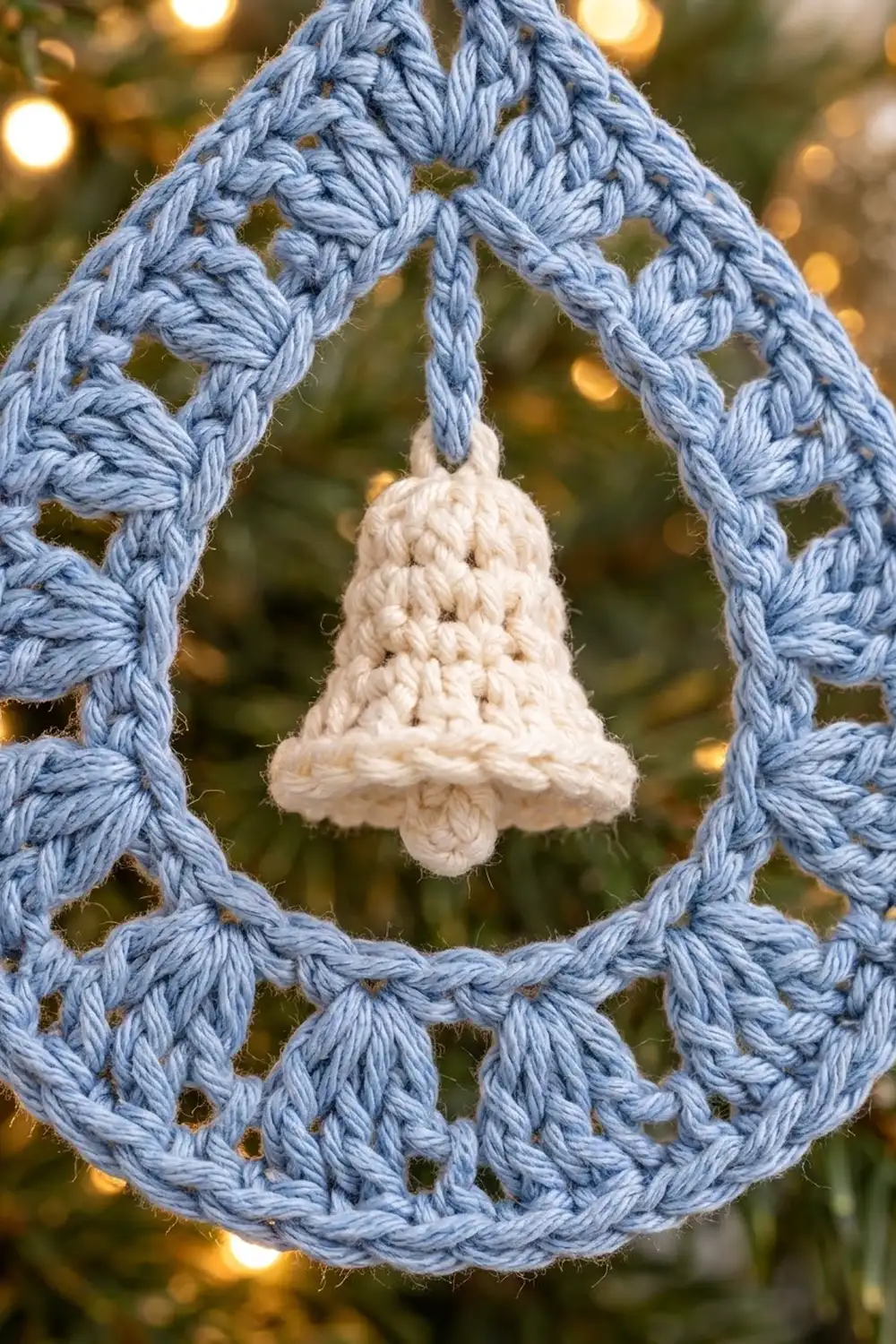 A white crocheted bell ornament suspended in a blue crochet diamond frame, with golden bokeh lights in the background.