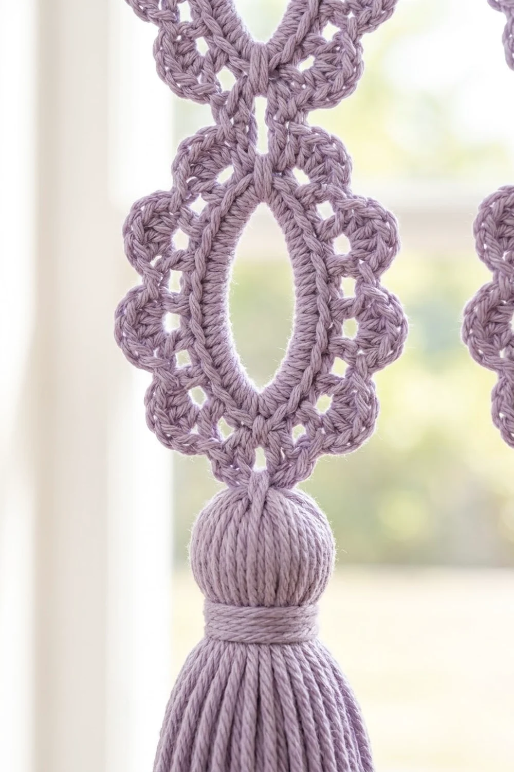 Lavender crocheted ornament with intricate openwork pattern and gathered tassel bottom, backlit by a blurred window.
