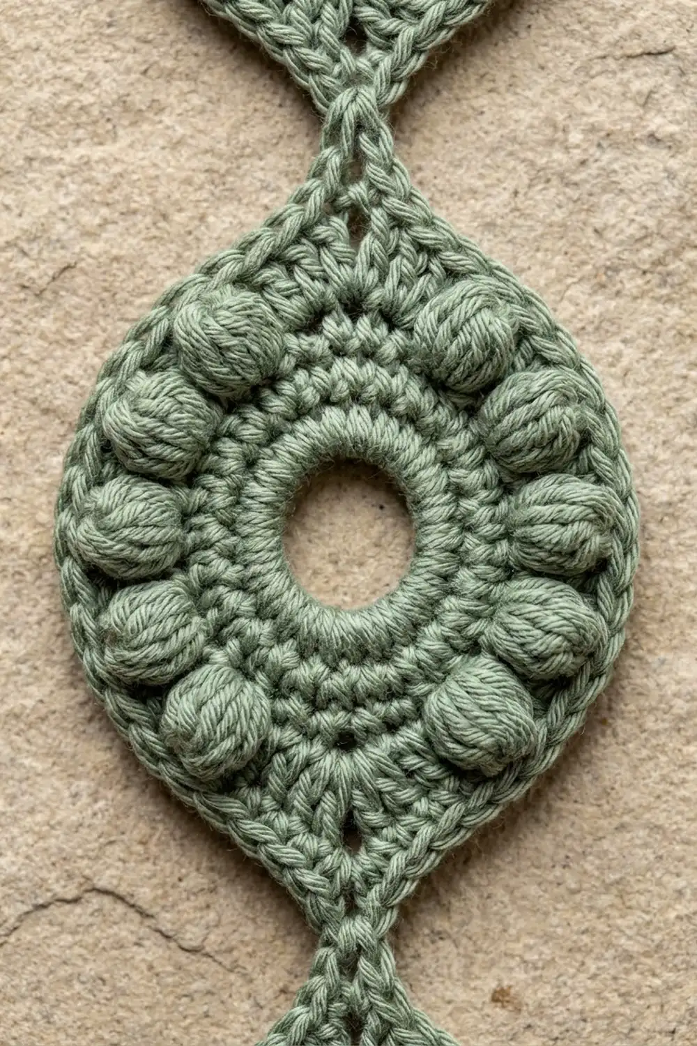 Sage green crocheted teardrop pendant with popcorn stitch detail photographed on neutral beige concrete.