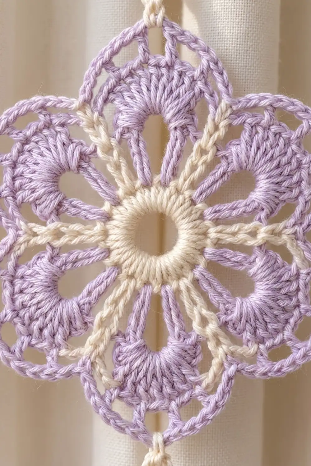 Delicate crochet doily in lavender and cream displayed as a hanging ornament against a neutral, textured background.