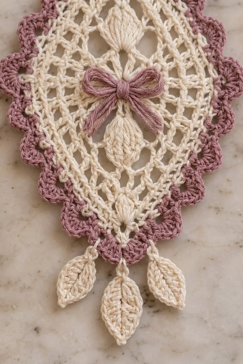 Cream and mauve crocheted potholder with openwork lace pattern, dusty pink bow, and leaf-shaped embellishments on marble.